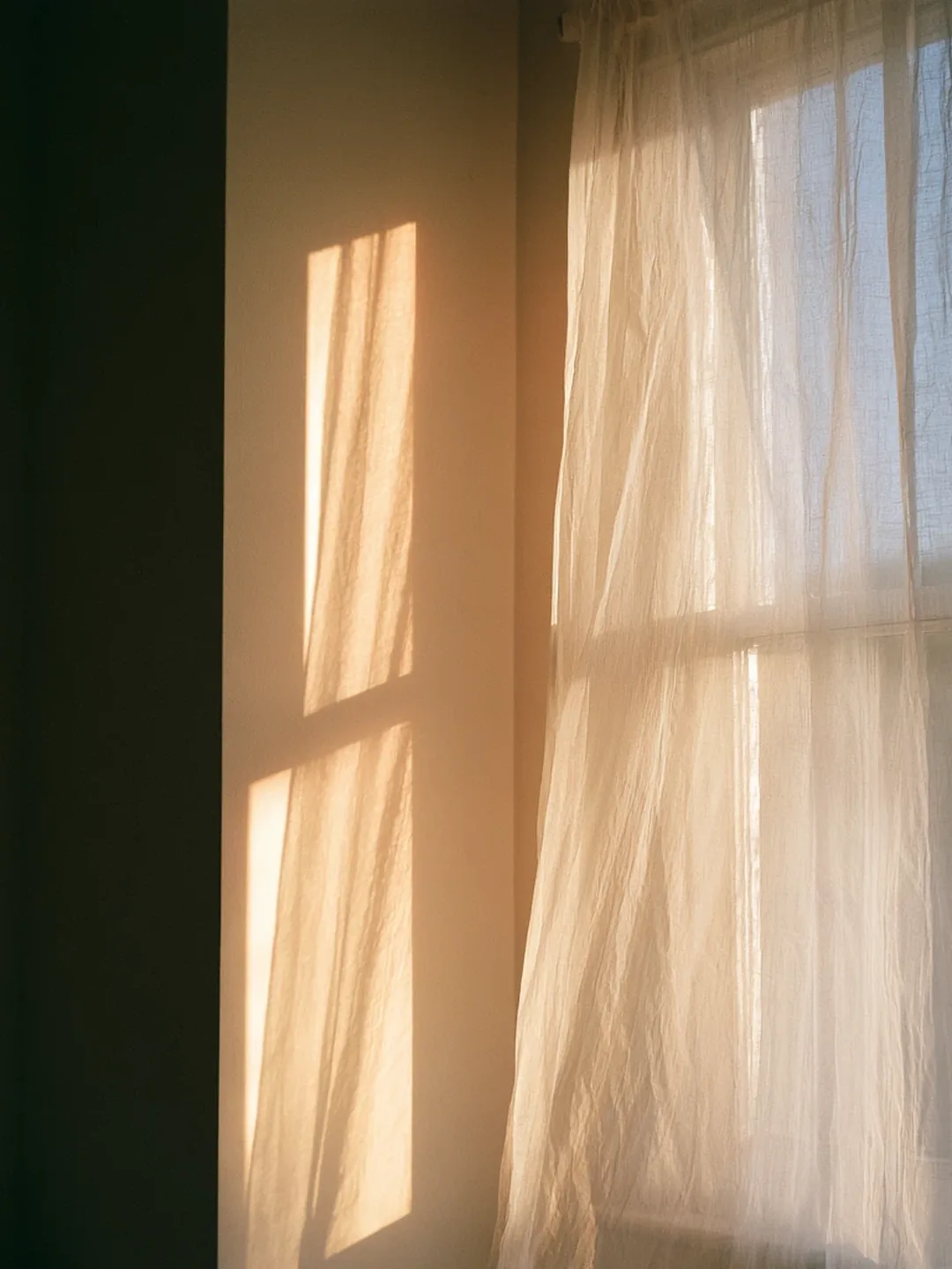 Late afternoon sunlight falling across a plain wall and a linen curtain in a quiet room.
