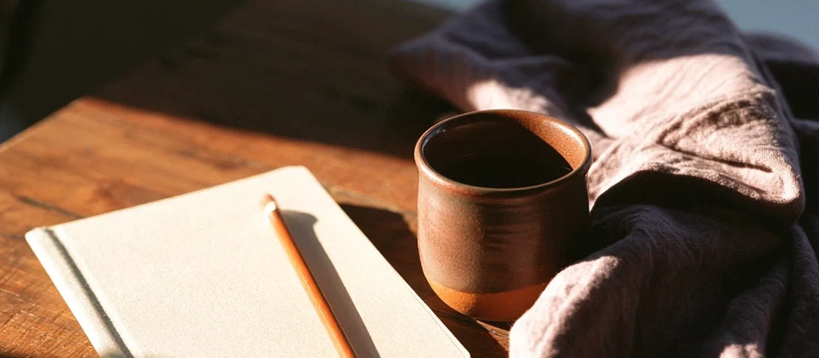 An open notebook and pencil beside a stoneware cup on a wooden table, lit by low sun.